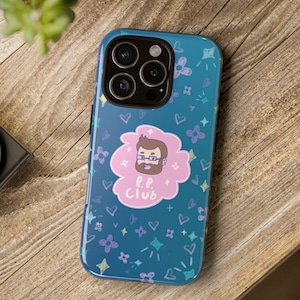 May include: A blue phone case with a cartoon illustration of a man with glasses and the text "P.P. Club" inside a pink cloud. The case is decorated with hearts, stars, and flowers. The phone case is on a wooden surface.
