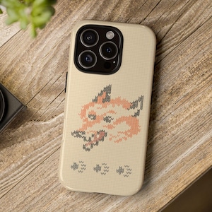 May include: A beige phone case with a knitted fox design in shades of peach, gray, and white. The case has a textured appearance and a camera cutout. The phone case is on a wooden surface.