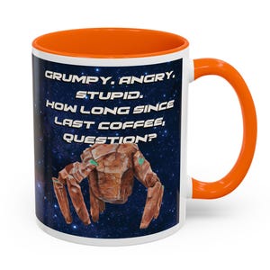 May include: White ceramic coffee mug with an orange interior and handle. The mug features a space-themed design with a brown creature and the text: "GRUMPY. ANGRY. STUPID. HOW LONG SINCE LAST COFFEE, QUESTION?"