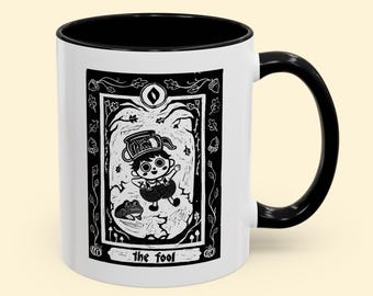 May include: A white ceramic mug with a black interior and handle. The mug features a black and white illustration of a character from the "Over the Garden Wall" cartoon, with the text "the fool" at the bottom.
