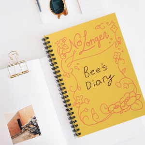 May include: A spiral-bound notebook with a yellow cover featuring the text "No Longer Bee's Diary" in a decorative font. The cover is adorned with floral illustrations in a coral color. The notebook is open, revealing blank pages.