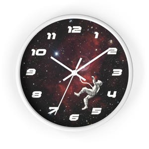 May include: White-framed wall clock with a black face featuring a space scene. The clock face displays an astronaut floating in space against a backdrop of stars and nebulae. White numerals and hands complete the design.