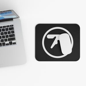 May include: A black rectangular mousepad with a white graphic of a stylized hand and a white computer mouse. A silver laptop is partially visible in the upper left corner, set against a white background.