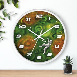 May include: White-framed wall clock with a space-themed design. The clock face features a green and orange nebula background, white numerals, and a silver astronaut. The clock hands are white.