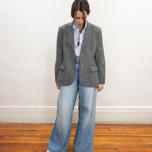 May include: A gray blazer with a button closure and a light blue striped shirt underneath. The person is wearing wide-leg blue jeans.