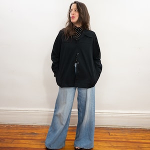 May include: A woman wearing a black button-up shirt over a black and white polka dot shirt and blue wide-leg jeans.