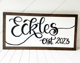 May include: A white wooden sign with a brown frame. The sign has black cursive lettering that reads "Eckles est. 2023".
