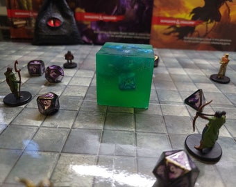 May include: A green, translucent cube with a blue liquid inside. The cube is surrounded by miniature figures and dice on a gray and white tiled surface.