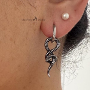 May include: A silver hoop earring with a detailed snake pendant. The snake design features textured scales and a curved body. A small pearl stud earring is also visible. The earrings are worn on a person's ear.