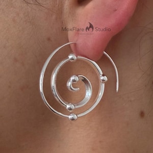 Pode incluir: Brincos espirais prateados com três pequenos detalhes de bolas. Os brincos têm um design único e moderno. São mostrados em close-up, destacando os detalhes intrincados e o acabamento polido.