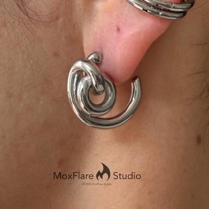 May include: Close-up of a silver-toned earring with a knot design and a matching ear cuff. The earring has a spiral shape and is worn on the ear. The ear cuff has a layered design. The image includes the text "MoxFlare Studio".