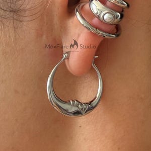 Puede incluir: Primer plano de una oreja adornada con pendientes de plata. Un pendiente de aro en forma de media luna cuelga del lóbulo, mientras que tres puños apilados con un acento de perla están en la parte superior de la oreja. El logotipo de MoxFlare Studio es visible.
