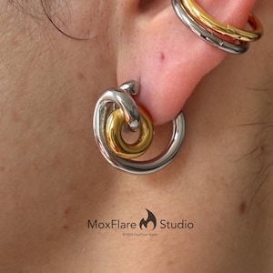 May include: Close-up of an ear with two sets of earrings. One earring is a silver hoop with a gold knot design. The other is a gold and silver ear cuff. The image is from MoxFlare Studio.
