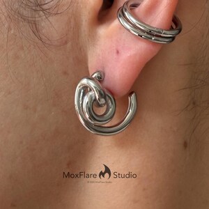 May include: Silver-toned ear jewelry set. The set includes a double-banded ear cuff and a large, swirling earring. The earring features a knot design at the top and a curved, spiral shape. The jewelry is from MoxFlare Studio.