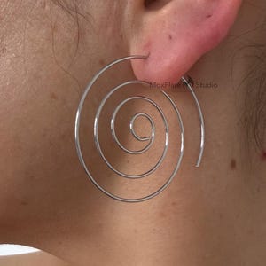 Puede incluir: Primer plano de un pendiente de espiral plateado. El pendiente es una línea continua que forma una espiral. El pendiente se lleva en la oreja. El fondo es la piel de una persona.