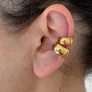 May include: A gold-toned ear cuff featuring a double-band design with a smooth, polished finish. The jewelry piece consists of two stacked, rounded bands that wrap around the cartilage of the ear, creating a layered appearance with reflective surfaces.