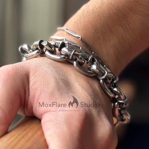 May include: A silver-colored chain bracelet with large, rounded links is worn on a wrist. A second, thinner bracelet is also visible. The bracelets are made of a shiny metal. The hand is holding a wooden object.