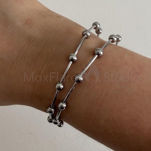 Pode incluir: Uma pulseira de metal prateado com uma série de pequenas contas esféricas espaçadas uniformemente ao longo de um fio fino e flexível. O design consiste em duas voltas de fio com contas, criando uma aparência em camadas com acabamento polido.