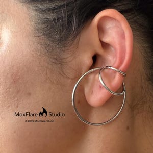 May include: A silver ear cuff featuring a large hoop and two smaller connected hoops. The ear cuff is worn on the ear. The MoxFlare Studio logo is visible in the lower left corner.