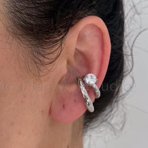 May include: A silver-toned ear cuff featuring a smooth, polished band and a second band adorned with a single, round, clear faceted stone. The jewelry is positioned on the cartilage of the ear, with the text MoxFlare Studio visible.
