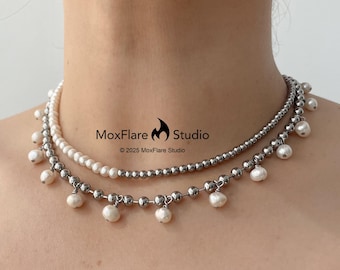 May include: Two layered necklaces. The top necklace is a strand of small white pearls. The bottom necklace is made of small silver beads with dangling white pearls. The necklaces are displayed on a person's neck. The image includes the text "MoxFlare Studio ©2025 MoxFlare Studio".