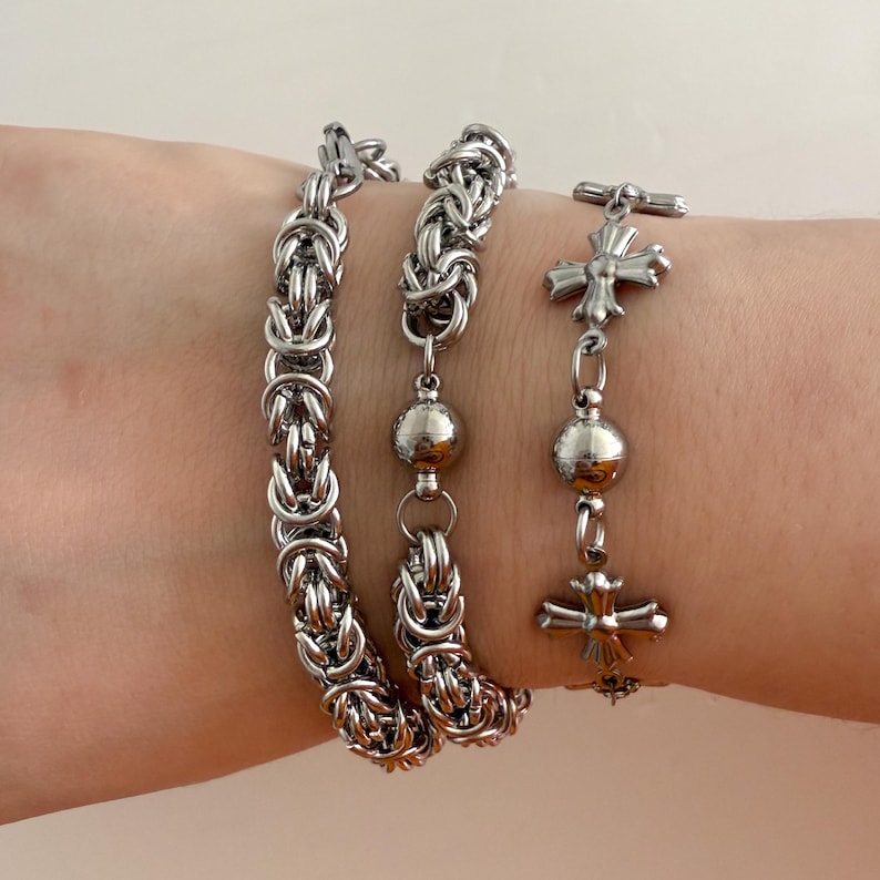 May include: Three silver bracelets on a wrist. The bracelets are made of chain links and one has a silver cross charm and silver beads.