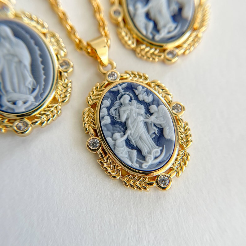 May include: Gold chain necklace with a blue and white cameo pendant featuring a religious figure. The pendant is surrounded by a gold leaf design and has small clear stones.