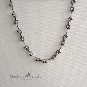 May include: A silver-toned necklace with a delicate design. The necklace features a series of small, round beads connected by curved, bar-shaped links. The MoxFlare Studio logo is visible in the bottom left corner.