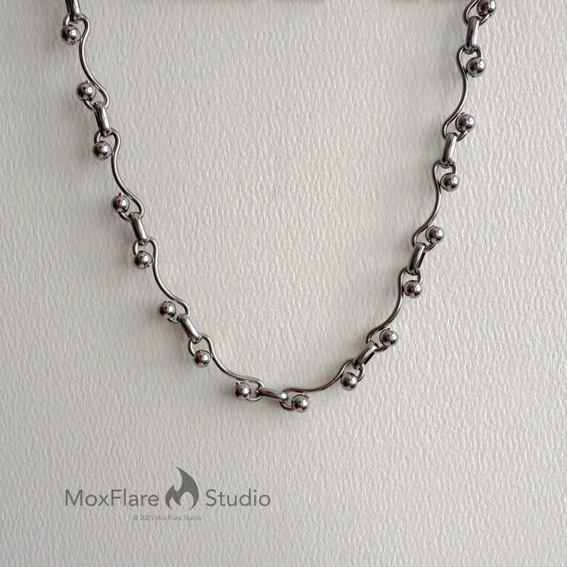 May include: A silver-toned necklace with a delicate design. The necklace features a series of small, round beads connected by curved, bar-shaped links. The MoxFlare Studio logo is visible in the bottom left corner.