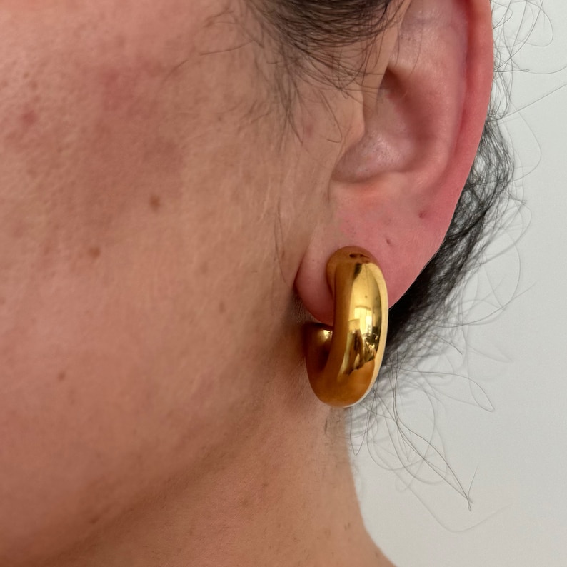 May include: Gold hoop earrings with a thick, rounded design. The earrings are worn in a person's ear.