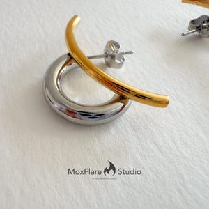May include: A pair of modern earrings. One earring features a silver-colored, circular base with a gold-colored, curved bar crossing over it. The earrings have a post-and-butterfly-clasp closure. The MoxFlare Studio logo is visible.