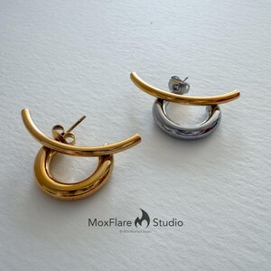 May include: Two modern earrings. One is gold-toned, the other silver-toned. Each earring features a curved bar above a circular hoop. The earrings are displayed on a white surface. The MoxFlare Studio logo is visible.