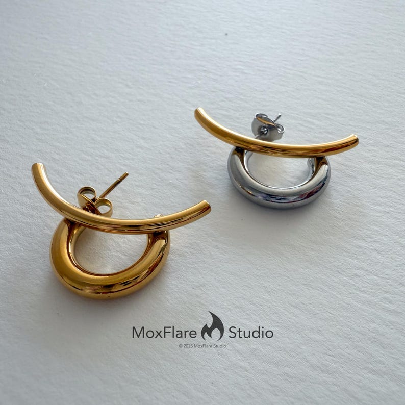 May include: Two modern earrings. One is gold-toned, the other silver-toned. Each earring features a curved bar above a circular hoop. The earrings are displayed on a white surface. The MoxFlare Studio logo is visible.