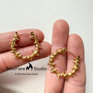 May include: A pair of gold-colored hoop earrings. Each earring features a series of outward-facing, pointed spikes around the hoop. The earrings are held up by a person's fingers. The MoxFlare Studio logo and copyright information are visible.