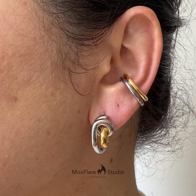 May include: A silver and gold-toned ear cuff and earring set. The ear cuff has a layered design, with a gold band and a silver band. The earring is a silver and gold-toned stud with a unique, intertwined design. The jewelry is worn on the ear.