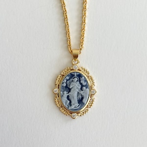 May include: Gold chain necklace with a blue and white cameo pendant featuring a religious figure surrounded by angels. The pendant is set in a gold frame with clear rhinestones.