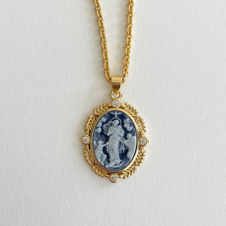 May include: Gold chain necklace with a blue and white cameo pendant featuring a religious figure surrounded by angels. The pendant is set in a gold frame with clear rhinestones.
