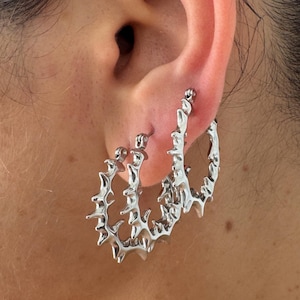 K&ouml;nnte beinhalten: Silberfarbene Creolen mit einem stacheligen, unregelm&auml;&szlig;igen Design. Die Ohrringe werden an einem Ohr getragen, wodurch ihr einzigartiges, strukturiertes Aussehen hervorgehoben wird. Die Creolen haben eine moderne, kantige &Auml;sthetik.