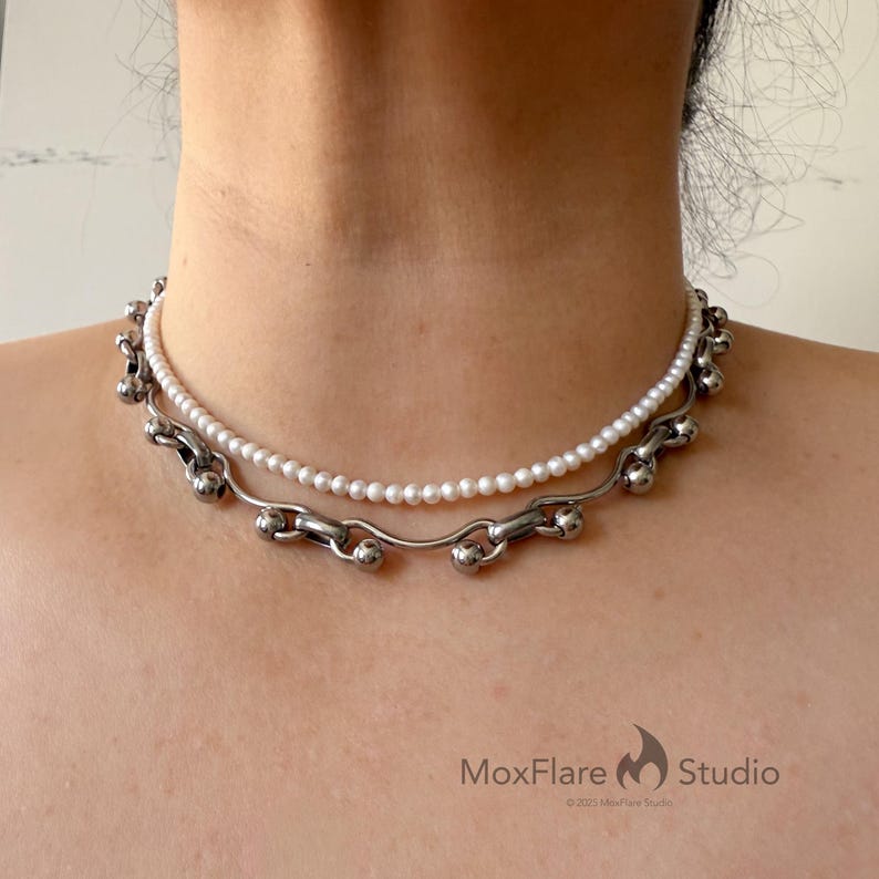 May include: A two-strand necklace featuring a delicate pearl strand paired with a silver-toned chain. The lower chain has a unique design with curved links and spherical accents. The necklace is displayed on a person's neck.