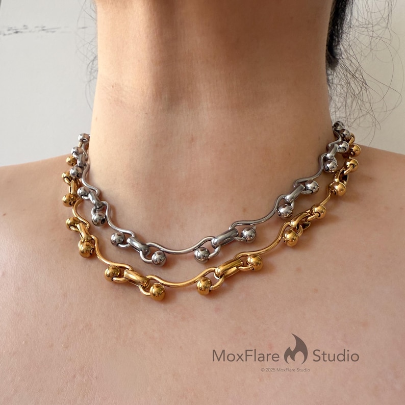 May include: Two necklaces, one silver-toned and one gold-toned, are displayed around a person's neck. The necklaces feature a chain-like design with rounded accents. The gold necklace is positioned below the silver one. The MoxFlare Studio logo is visible.