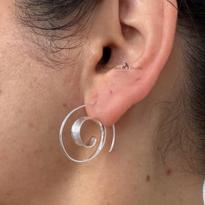 May include: A silver spiral earring is shown in close-up. The earring has a continuous, open spiral design with a textured centre. The earring is worn on an ear, with the ear and surrounding skin visible. The earring is a piece of jewellery.