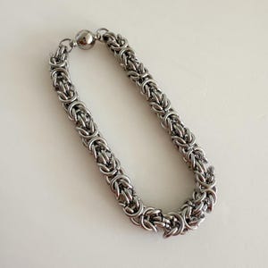 Pu&ograve; includere: Un braccialetto a catena argentato con chiusura magnetica. La catena &egrave; realizzata con anelli di metallo intrecciati.