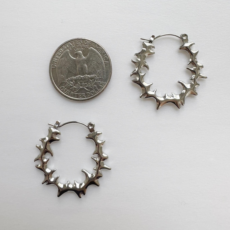 K&ouml;nnte beinhalten: Zwei silberfarbene Creolen mit stacheligem Design, neben einer US-Vierteldollarm&uuml;nze. Die Ohrringe haben eine runde Form mit nach au&szlig;en gerichteten Spitzen, die einen auff&auml;lligen Look erzeugen. Durchmesser ca. 2,5 cm.