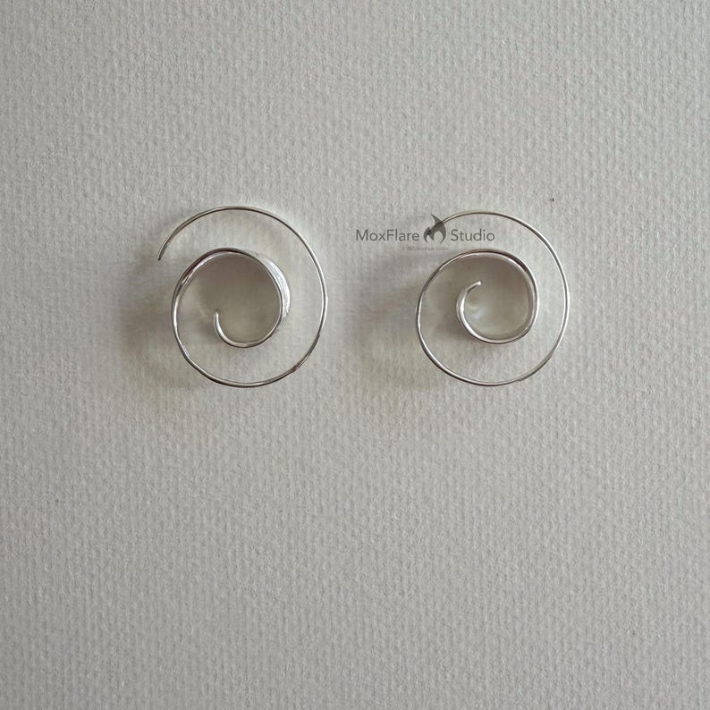 May include: A pair of silver spiral earrings. The earrings are in a spiral shape, with a smooth, polished finish. The earrings are displayed on a light grey background. The earrings are from MoxFlare Studio.