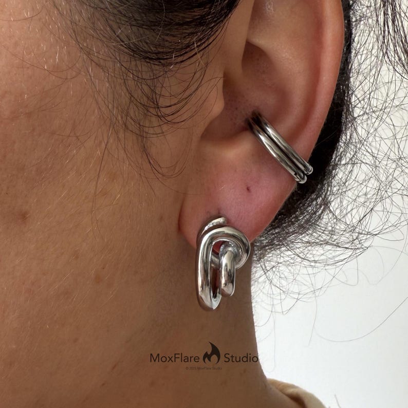 May include: Silver-toned ear cuff and earrings set. The ear cuff features a double-band design, while the earrings have a looped, abstract shape. The jewelry is worn on a person's ear, with the focus on the metallic accessories.