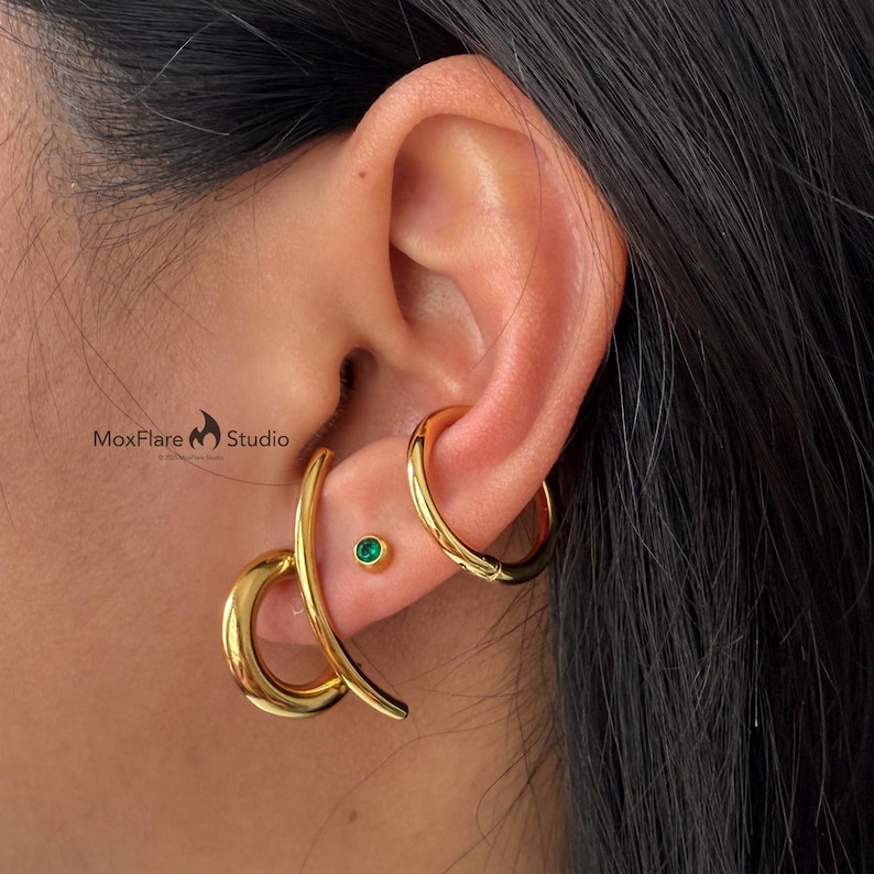 May include: Close-up of an ear adorned with gold earrings. The ear features a small, round, emerald-green stud and two larger gold earrings: a curved, elongated design and a classic hoop. The image is branded with "MoxFlare Studio".