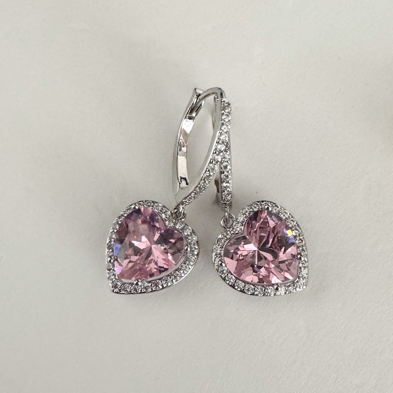 May include: A pair of silver dangle earrings with pink heart-shaped stones surrounded by a halo of smaller clear stones.