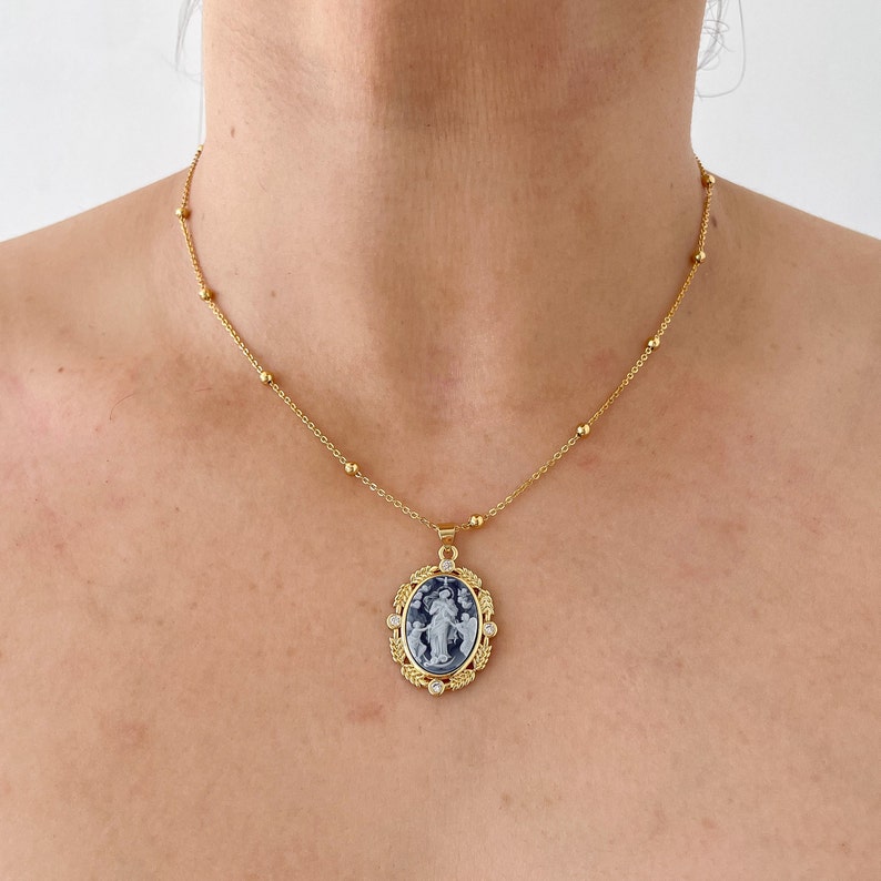 May include: A gold chain necklace with a blue and white oval pendant. The pendant has a detailed design of a woman holding a baby. The pendant is surrounded by small gold beads and a gold frame.