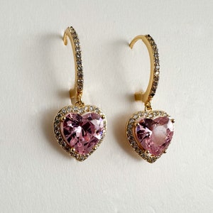 May include: A pair of gold hoop earrings with a pink heart-shaped gemstone surrounded by smaller clear gemstones.