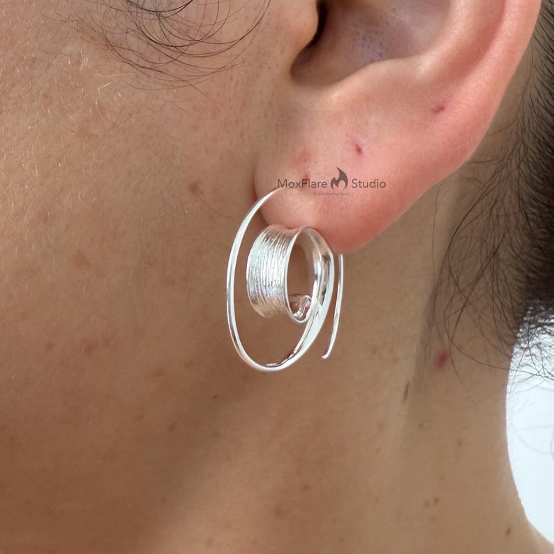 May include: A silver spiral earring is shown close up. The earring has a textured, cylindrical centre and a spiralling outer design. The earring is worn on an ear, highlighting its unique shape and metallic shine.